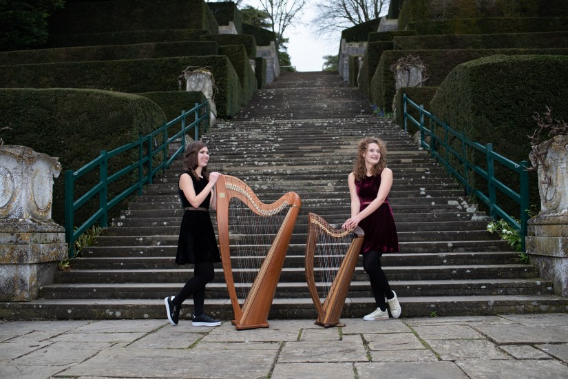 Two harpists standing by harps in front of staircase outdoors, left harp is bigger than the right harp. 